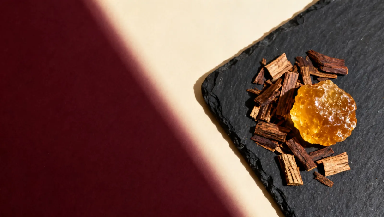 Natureza-morta em luz lateral dura: lascas de madeira de oud e um bloco de resina de ambar dourado, sobre fundo vinho e creme