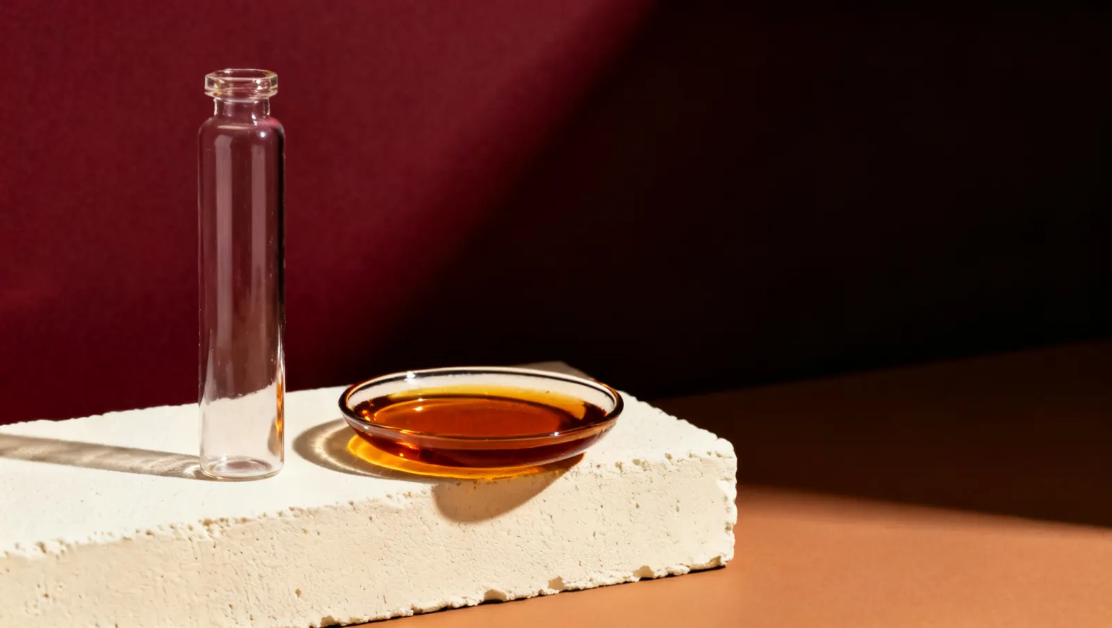 Natureza-morta em luz lateral dura: um frasco alto de vidro transparente e um prato raso com liquido ambar, sobre fundo vinho e creme