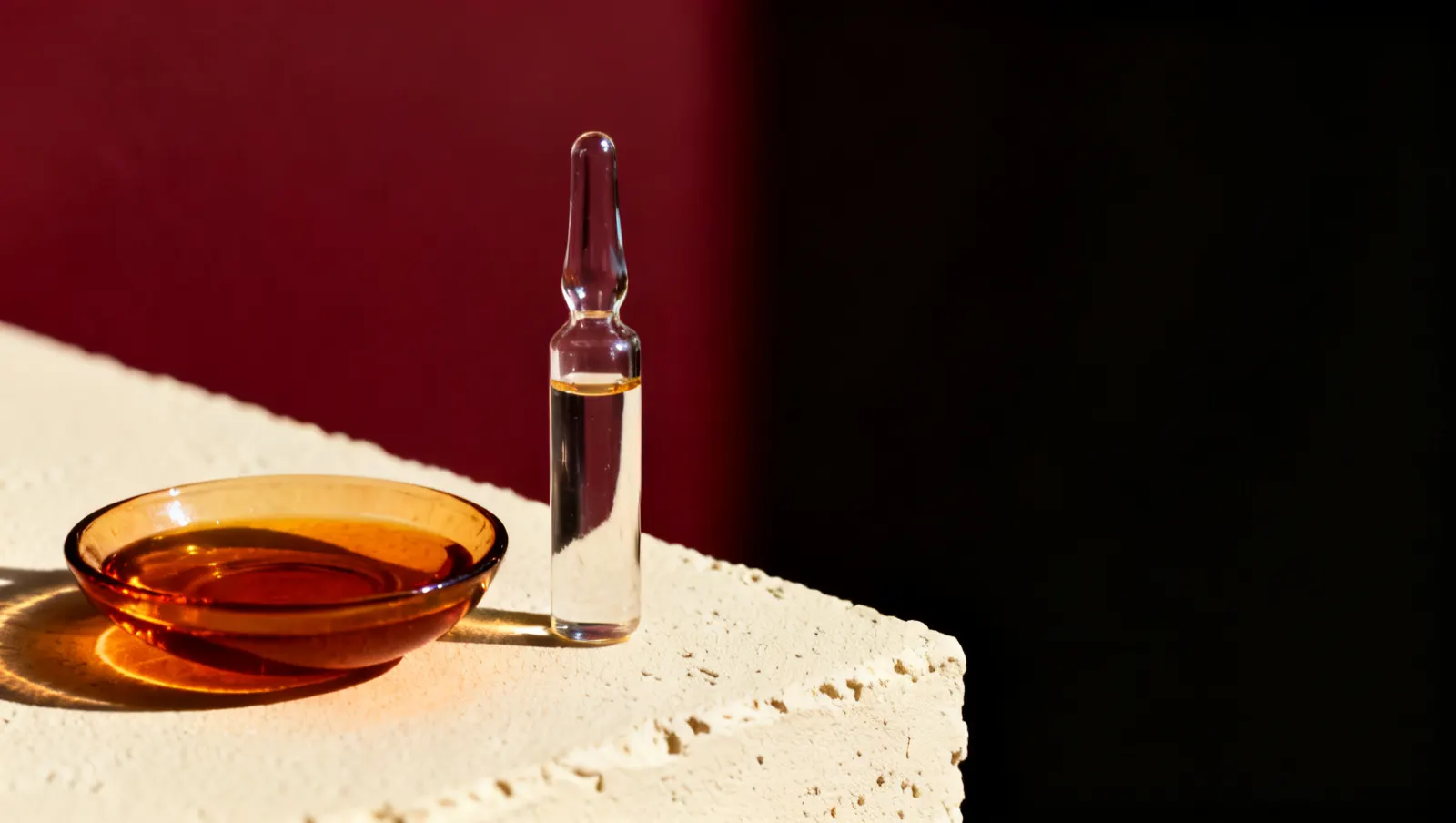 Natureza-morta em luz lateral dura: um prato raso com liquido ambar e um frasco alto de vidro transparente, sobre fundo vinho e creme