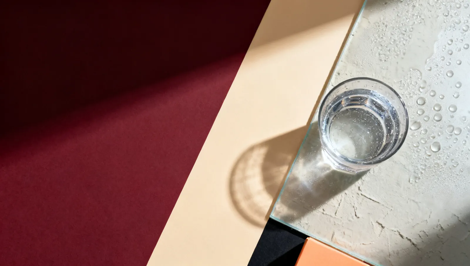 Natureza-morta em luz lateral dura: condensacao fria em gotas sobre vidro, sobre fundo vinho e creme