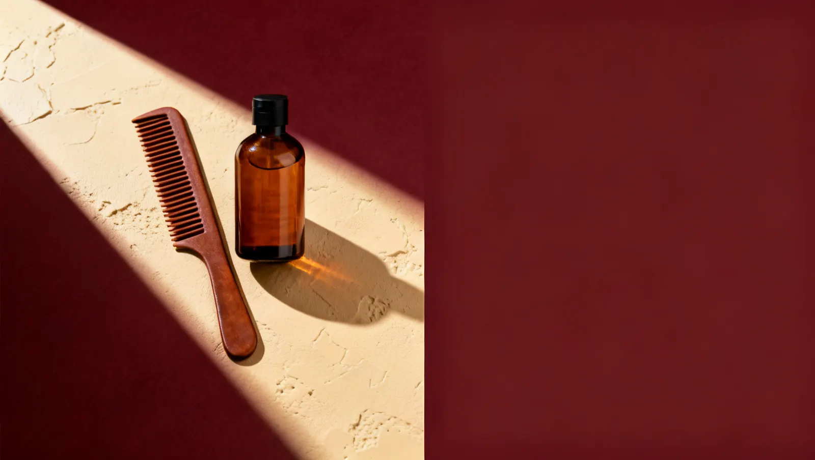 Natureza-morta em luz lateral dura: um pente de dentes largos e um frasco de vidro ambar sem rotulo, sobre fundo vinho e creme