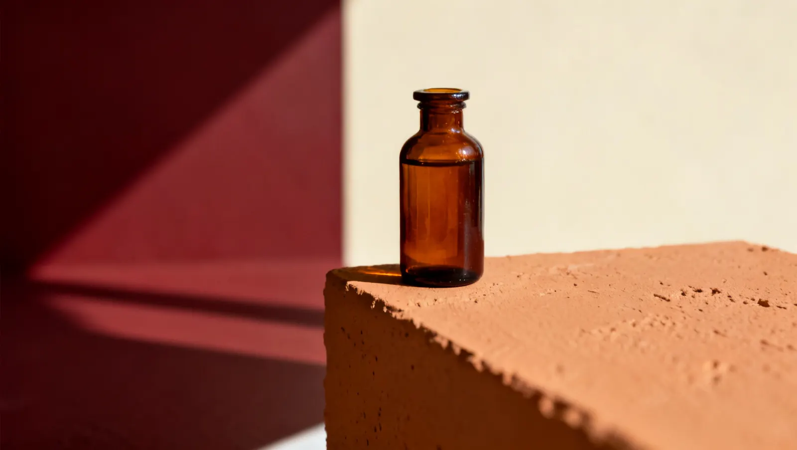 Natureza-morta em luz lateral dura: um frasco de vidro ambar sem rotulo, sobre fundo vinho e creme