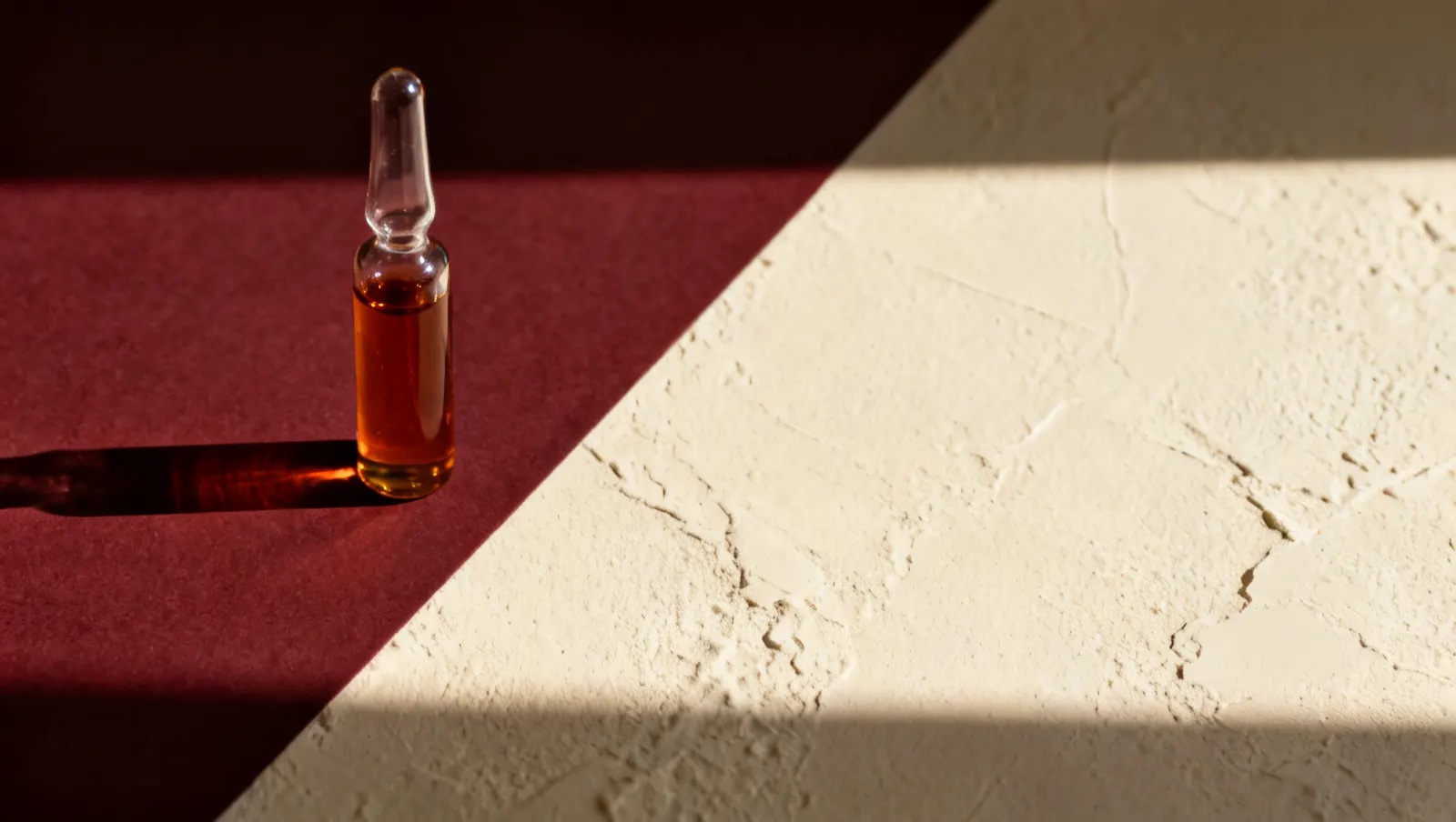 Natureza-morta em luz lateral dura: um frasco alto de vidro transparente, sobre fundo vinho e creme