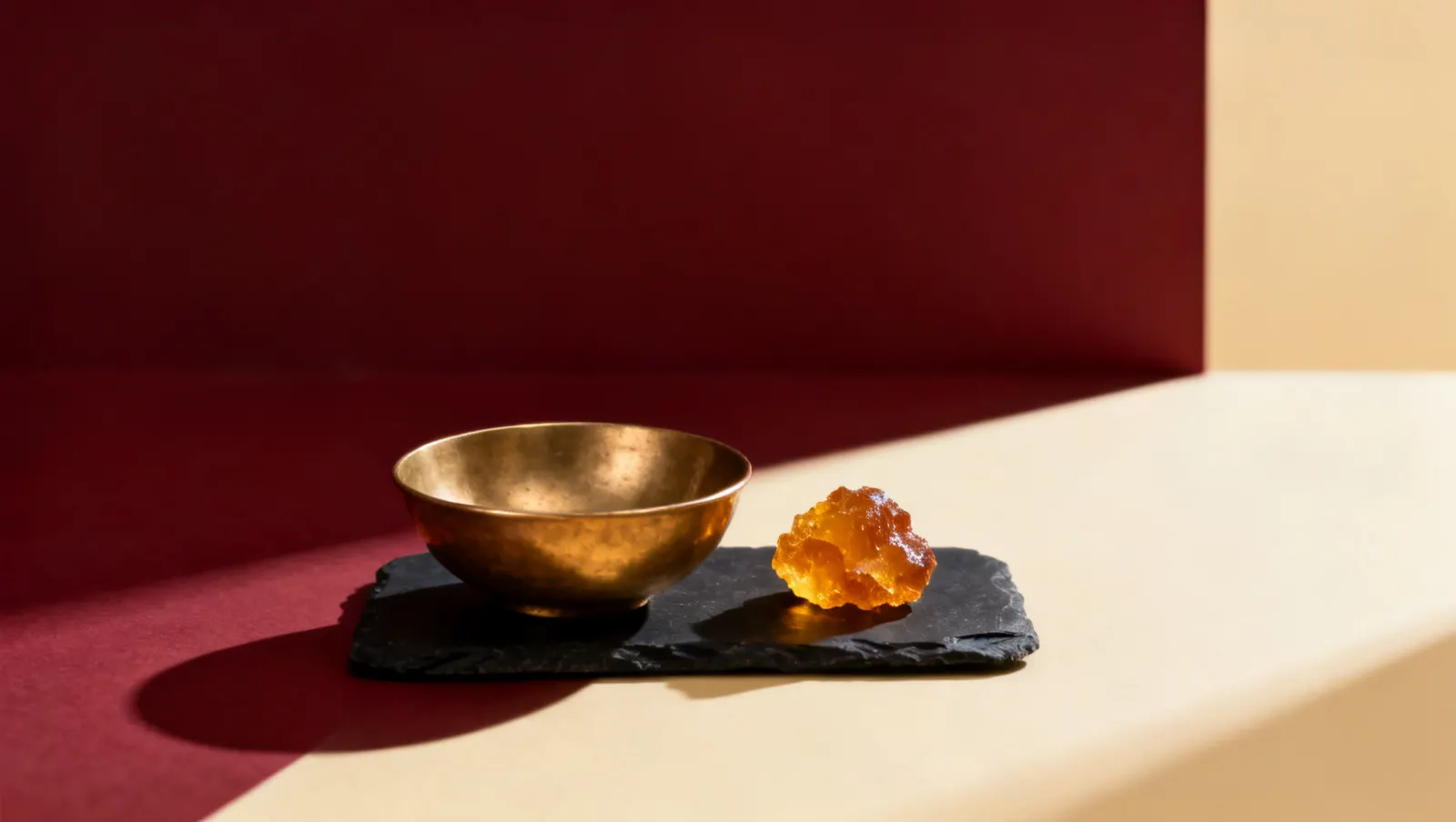 Natureza-morta em luz lateral dura: uma tigela lisa de latao e um bloco de resina de ambar dourado, sobre fundo vinho e creme