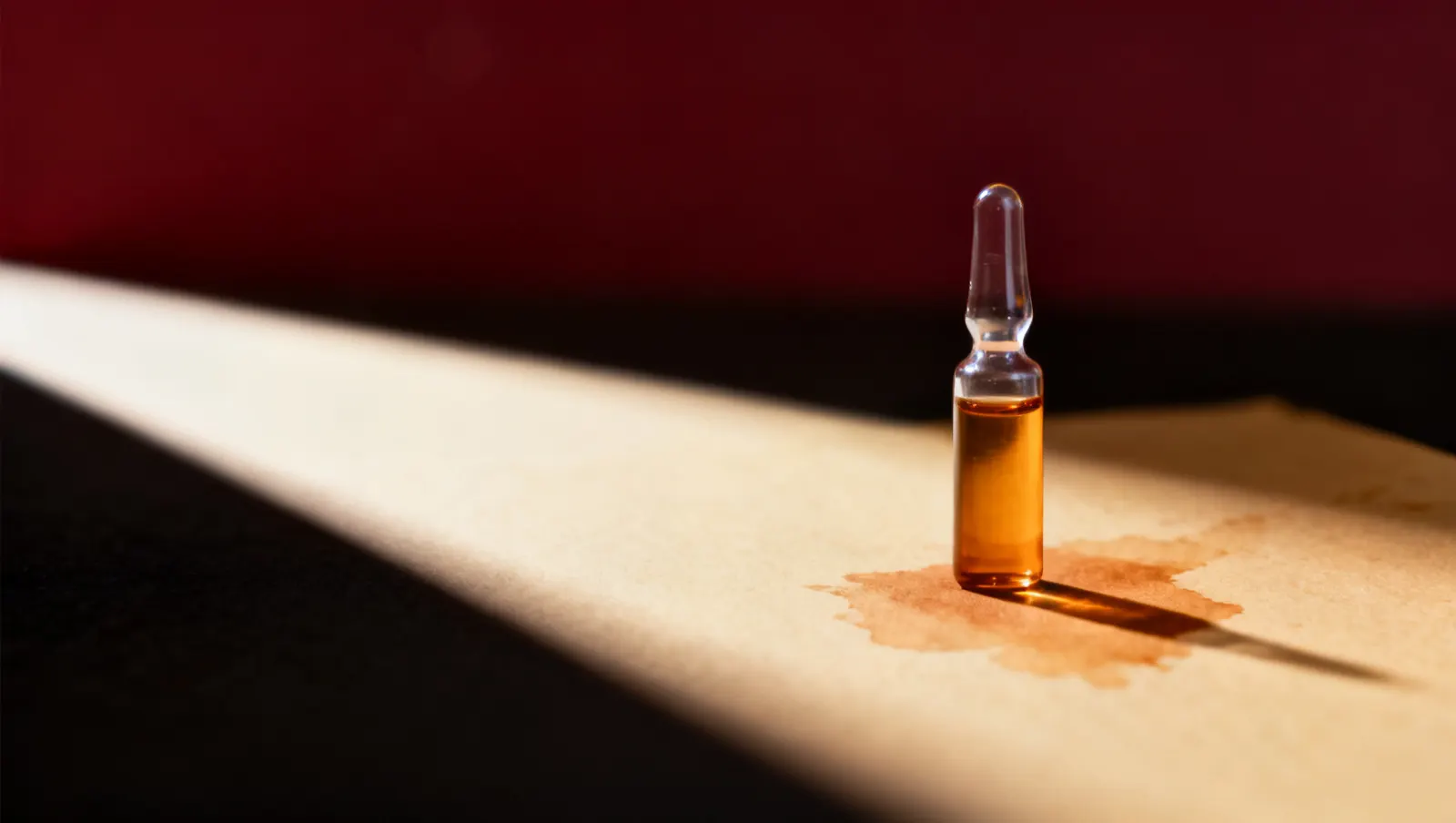 Natureza-morta em luz lateral dura: um frasco pequeno de vidro transparente, sobre fundo vinho e creme