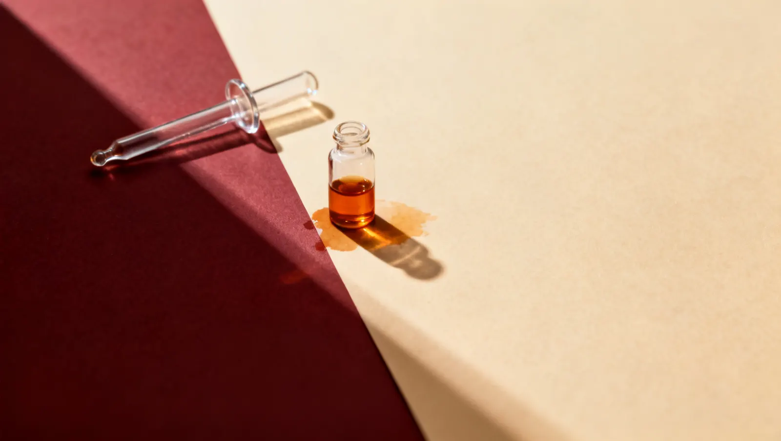 Natureza-morta em luz lateral dura: uma pipeta de vidro lisa e um frasco pequeno de vidro transparente, sobre fundo vinho e creme