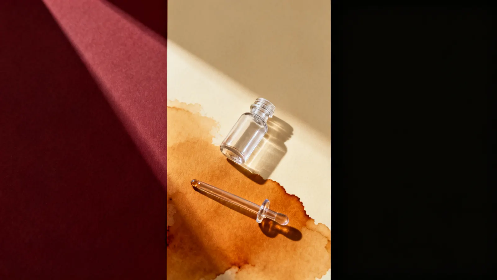 Natureza-morta em luz lateral dura: um frasco pequeno de vidro transparente e uma pipeta de vidro lisa, sobre fundo vinho e creme