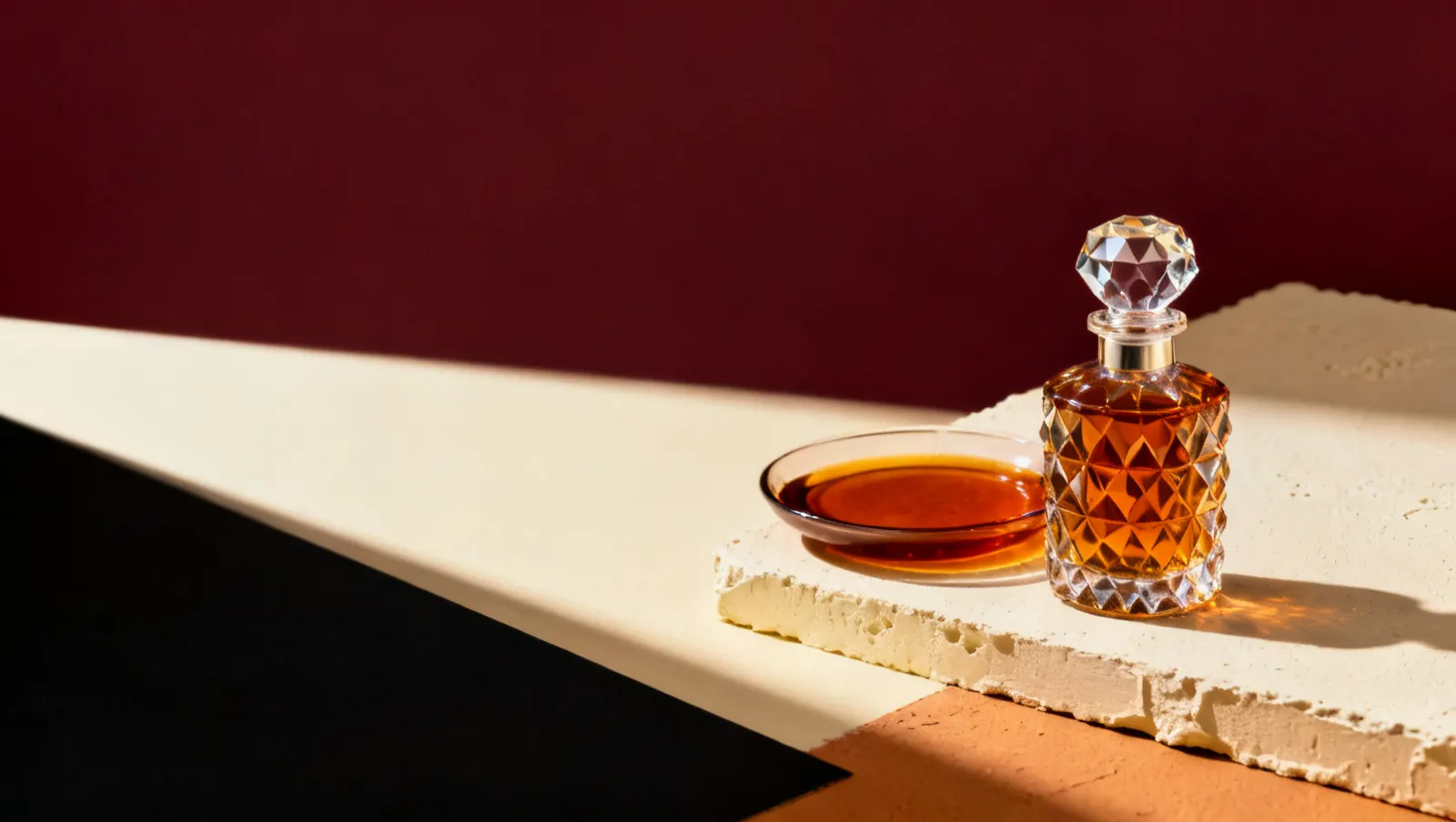 Natureza-morta em luz lateral dura: um prato raso com liquido ambar e um frasco de cristal facetado sem rotulo, sobre fundo vinho e creme