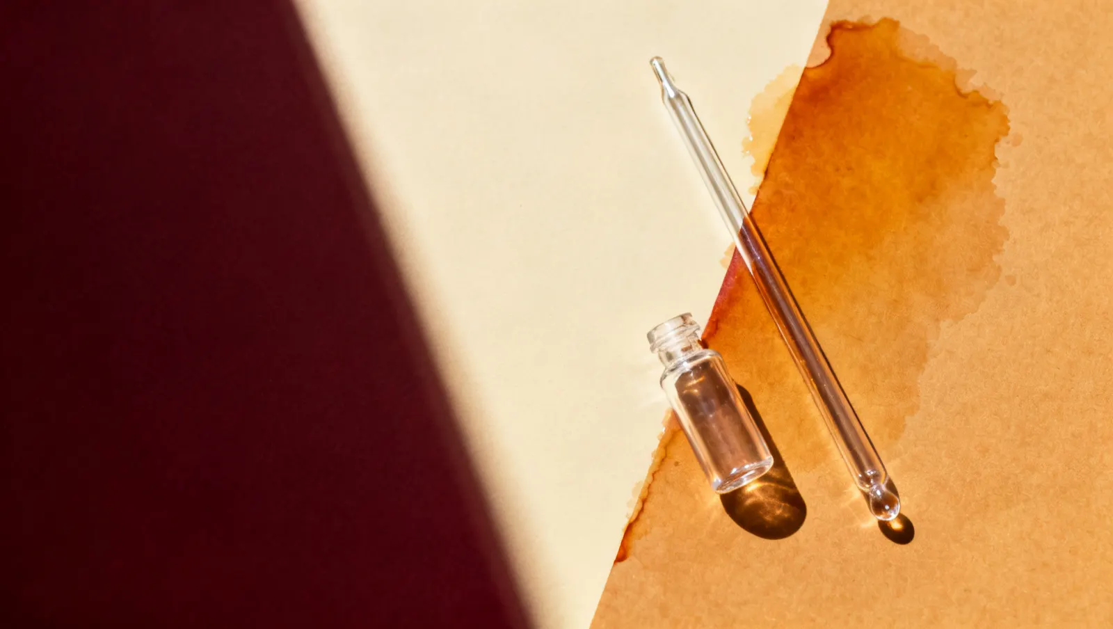 Natureza-morta em luz lateral dura: uma pipeta de vidro lisa e um frasco pequeno de vidro transparente, sobre fundo vinho e creme
