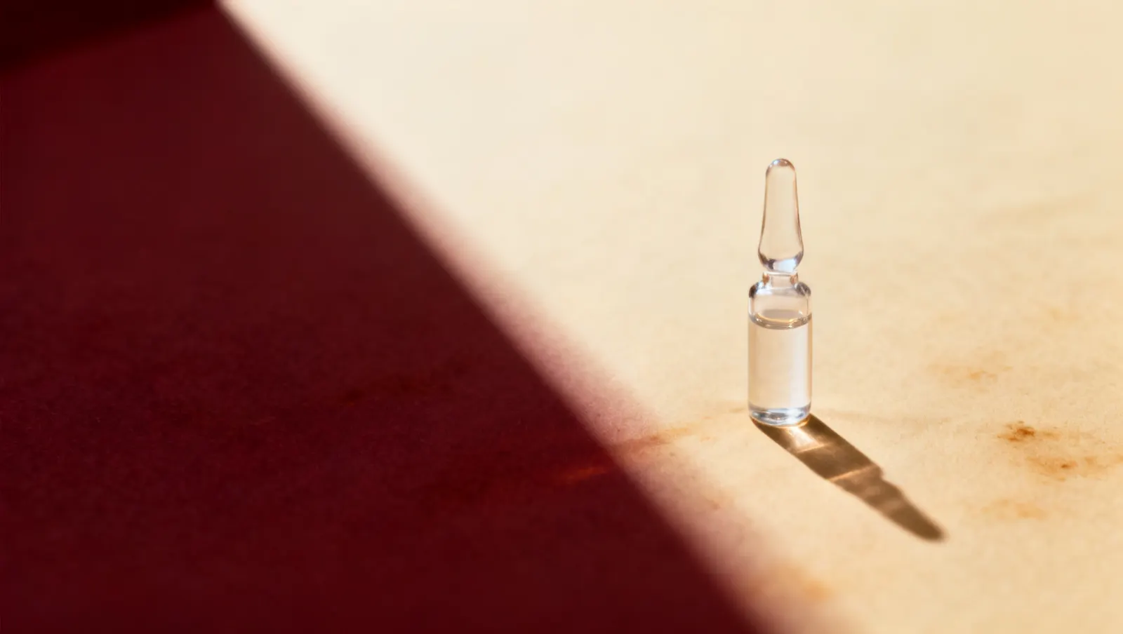 Natureza-morta em luz lateral dura: um frasco pequeno de vidro transparente, sobre fundo vinho e creme